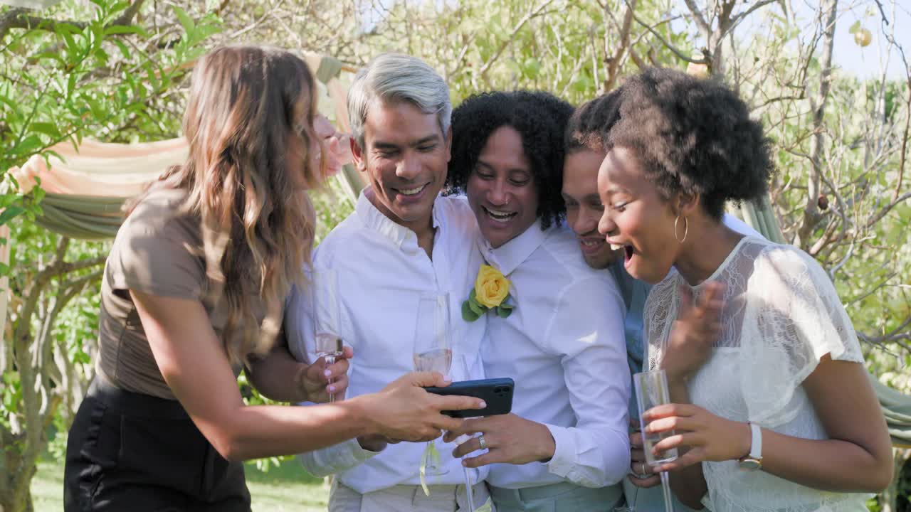 Five friends toasting with flutes in garden extending smartphone taking selfie showing boutonniere
