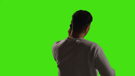 vista trasera de un joven respondiendo a una llamada en un teléfono móvil de pie contra el fondo de un estudio de pantalla verde con iluminación de baja clave