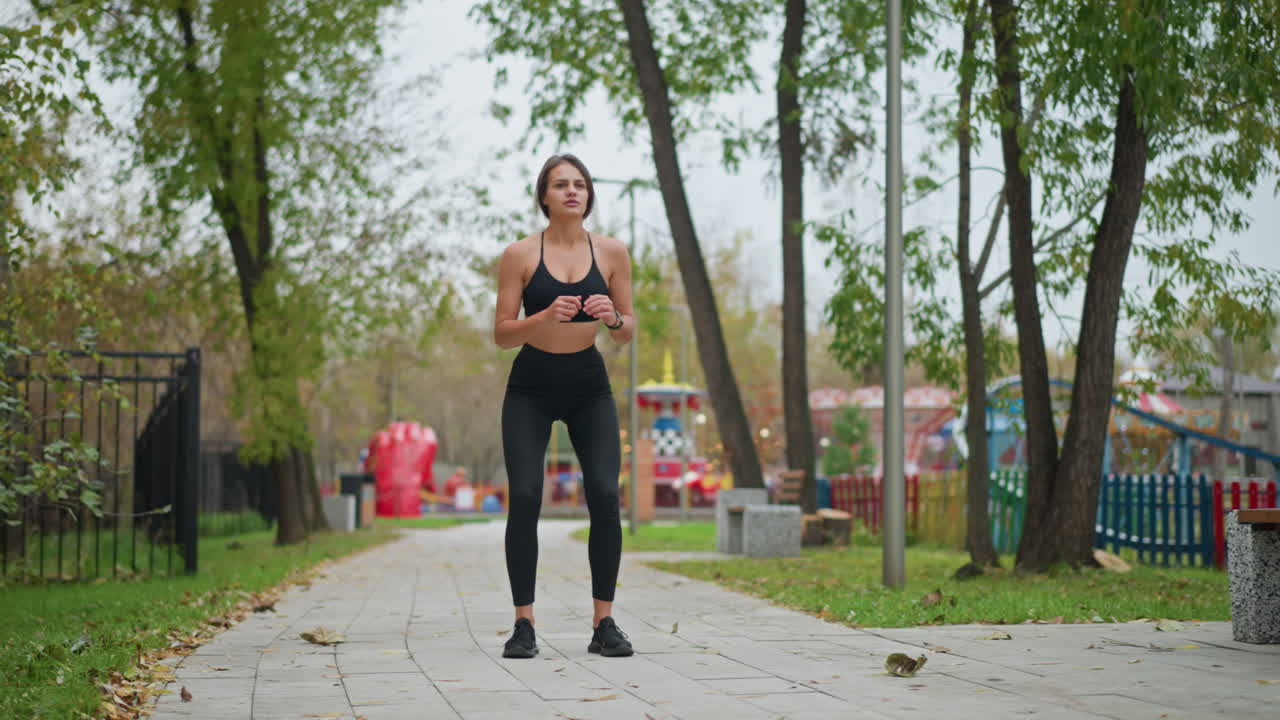 Young girl performing outdoor exercise in game park, squatting down, surrounded by trees and playground equipment, enhancing her fitness while enjoying outdoor activities