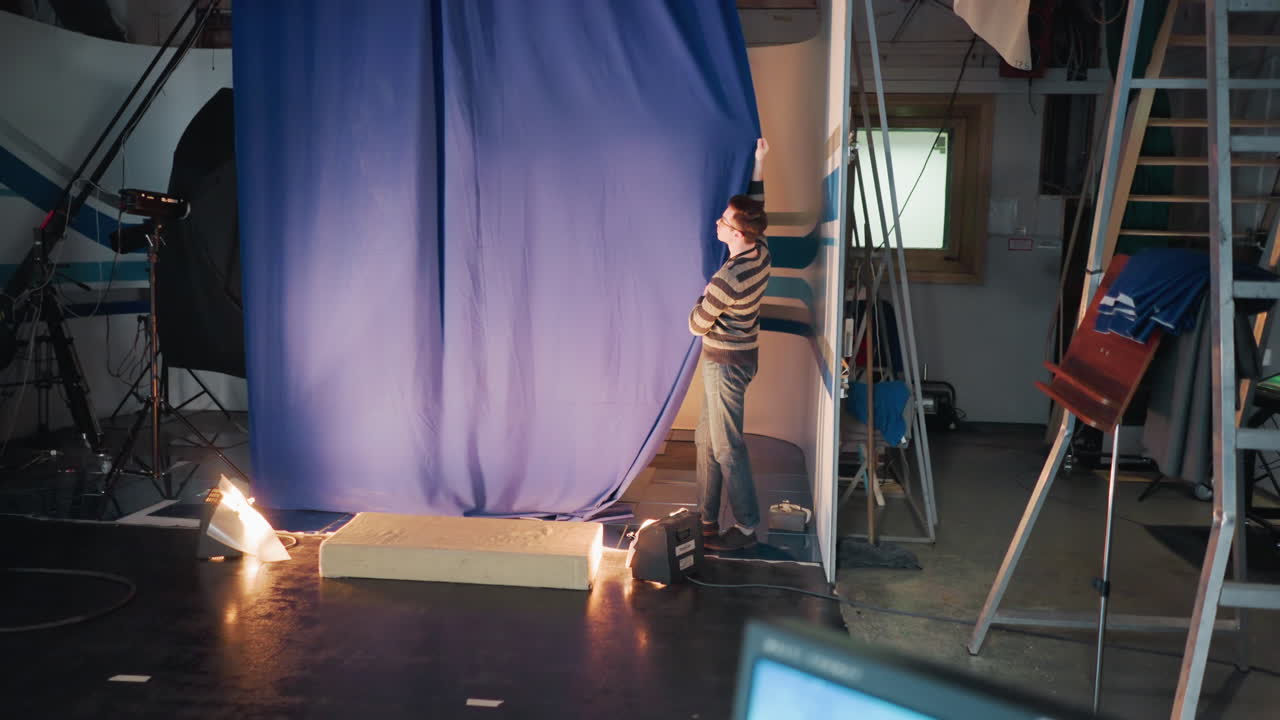 Studio crewman wearing striped sweater adjusts tall blue curtain during set preparation under lights among cameras cables ladders capturing scenes atmosphere of broadcast stage installation activity