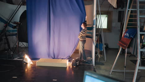 studio crewman wearing striped sweater adjusts tall blue curtain during set preparation under lights among cameras cables ladders capturing scenes atmosphere of broadcast stage installation activity