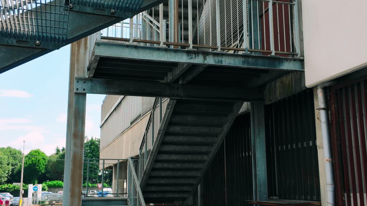 Tampilan geometris struktur tangga baja yang melekat pada bangunan modern di Bergamo. Arsitektur industri menonjolkan kisi-kisi logam, pagar, dan penopang struktural dengan latar belakang langit cerah.
