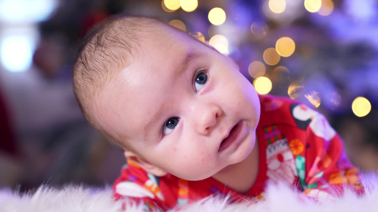 Adorable baby face with cute plump cheeks looks up. Sweet kid lies on a fluffy plaid close up. Christmas decorations in blur at backdrop.