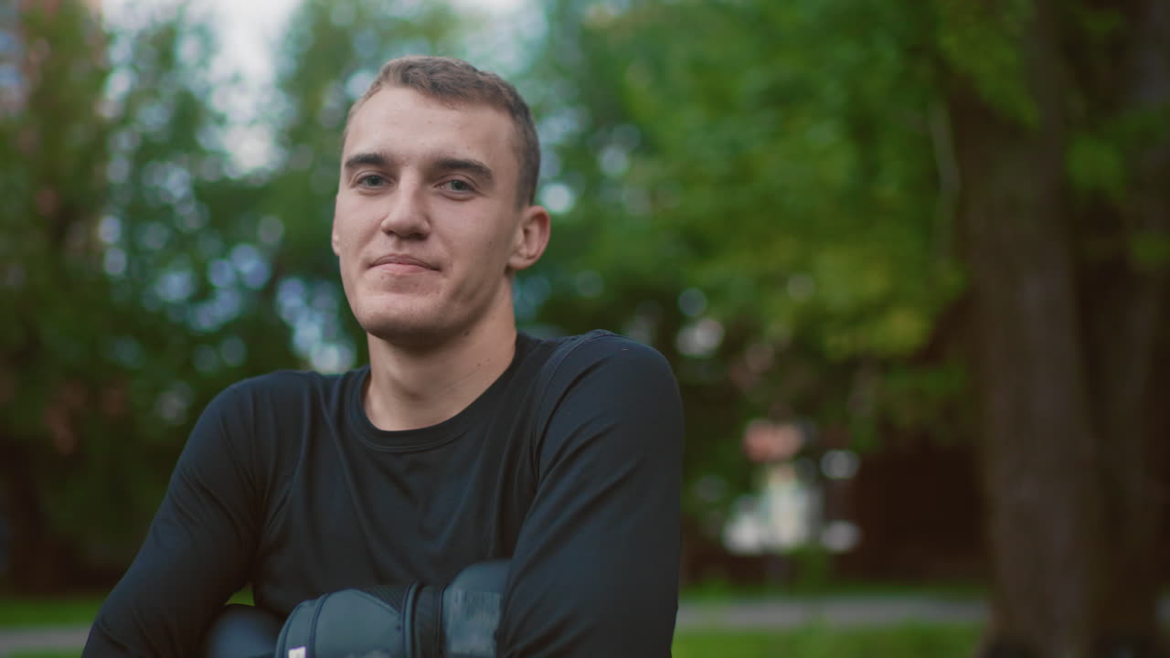 Ung, hvit mann med hansker, utendørs atletportrett av selvsikker bokser og trener iført svart, langermet trøye og polstrede hansker, armene i kryss, smilende og fokuserte uttrykk, parkbakgrunn med løv