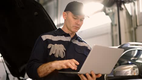 car mechanic working on laptop in auto repair service, lean on a car, low angle view