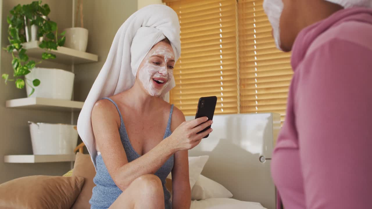 Amiche felici di diverse etnie con asciugamani in testa e maschere purificanti che usano lo smartphone a casa