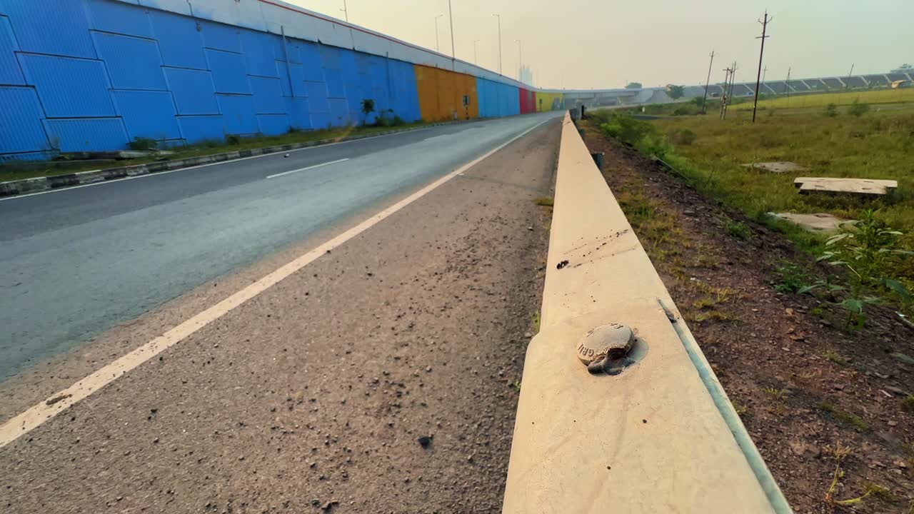 Tracking shot of a concrete guard rail running beside a highway overbridge, with colorful barriers and open fields in the background under soft daylight