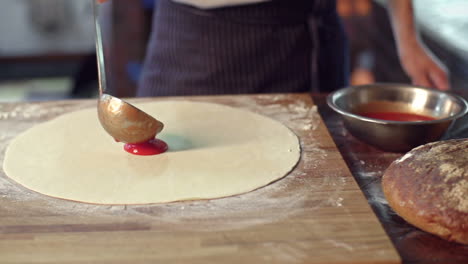 medio cuerpo de una mujer con un delantal, tomando una cacerola de salsa de tomate y vertiéndola sobre una masa delgada en la mesa de la cocina