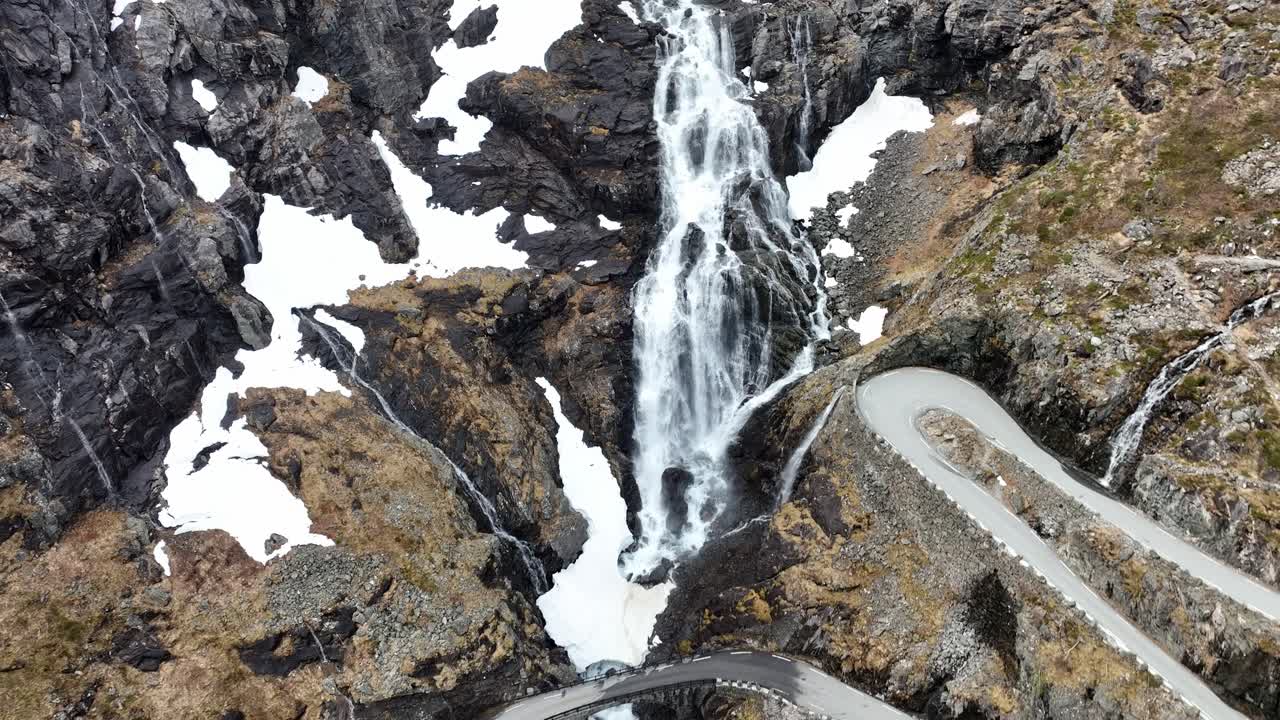aerial descendiendo por la cascada de stigfossen y revelando el puente - trollstigen sinuosa carretera de montaña en noruega durante la primavera en mayo con manchas de nieve todavía en el terreno