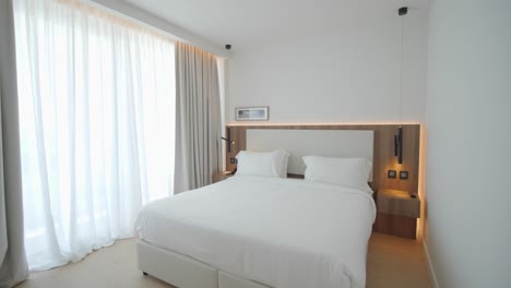 Una-Toma-Cardánica-De-Un-Dormitorio-Elegante-Dentro-De-Un-Apartamento-De-Hotel-Lleno-De-Luz-Con-Cortinas-Color-Crema-Y-Almohadas-Enormes-Y-Decoración-De-Madera-Con-Luces-LED