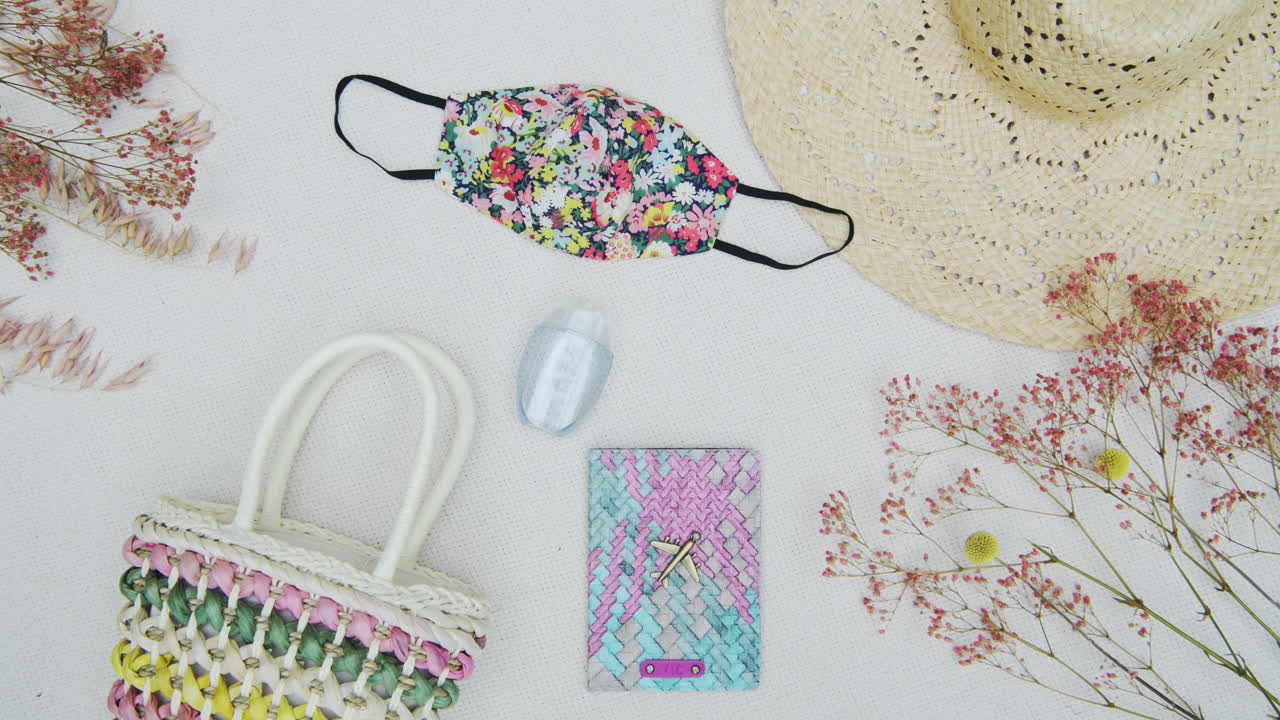 Panning through a minimalist view of modern essentials: face mask for coronavirus pandemic, a pastel-colored purse, with wide-brimmed sun hat against a pure white background and dried flowers