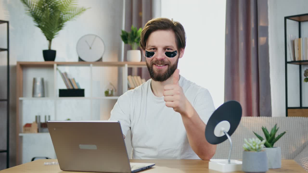 atractivo sonriente feliz joven moderno barbudo con colágeno parches negros para los ojos sentado frente a la computadora y mostrando el pulgar hacia la cámara