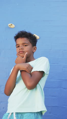 Vertical video: Posing confidently, child standing against blue wall with thoughtful expression