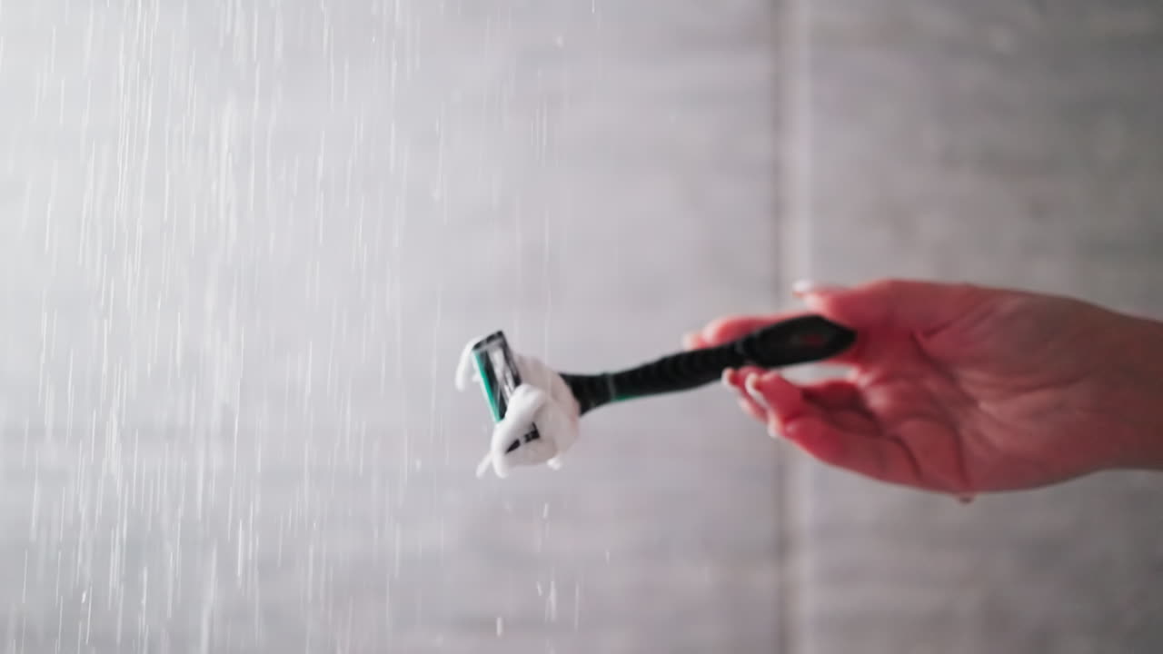 Woman cleans razor from foam in shower sprays closeup. Lady holds shaver under falling down water jets to wash out froth in bathroom. Shaving tool