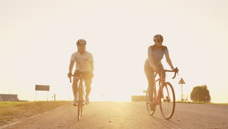 a man and a woman on bicycles ride down the road at sunset together in slow motion. the couple travels by bicycle. sports cycling helmets.