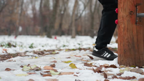 close-up of black sneakers on a snowy park path, focusing on leg movements as someone prepares to jump onto outdoor workout equipment, surrounded by fallen leaves and snow