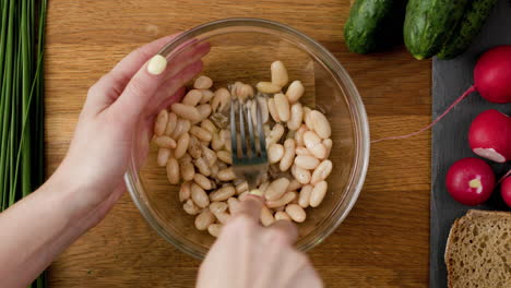 mashing white beans with a fork to prepare vegan sandwich spread with rye bread, seasoned with salt, pepper and olive oil