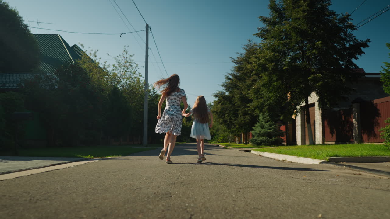 Rear view of woman in floral dress holding up skirt slightly as she runs hand in hand with young girl along quiet sunny residential street lined with green trees and houses on warm clear day