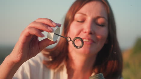 close up of fair skin woman gently blowing soapy bubble wand with rainbow reflections, eyelids lowered in peaceful expression, warm sunset light illuminating lips and hair