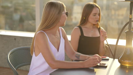 two beautiful girlfriends enjoying free time in the café outdoors. ladies talk, relax and smoking hookah in the rays of setting sun. blurred backdrop.