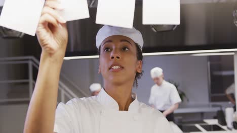 African-American-female-chef-working-in-a-restaurant-kitchen-checking-orders,-with-colleagues-workin