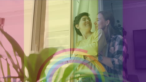 animation of pride rainbow flag and rings over happy caucasian lesbian couple embracing at home