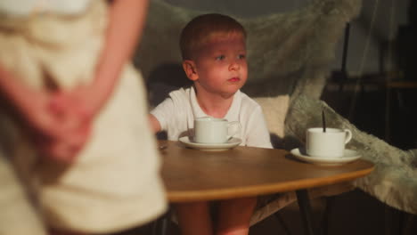 niño pequeño se sienta en la mesa de café con tazas de té y platillos. niño tranquilo descansa bebiendo bebida con la hermana en casa. niño adorable se inclina hacia el podio en la habitación