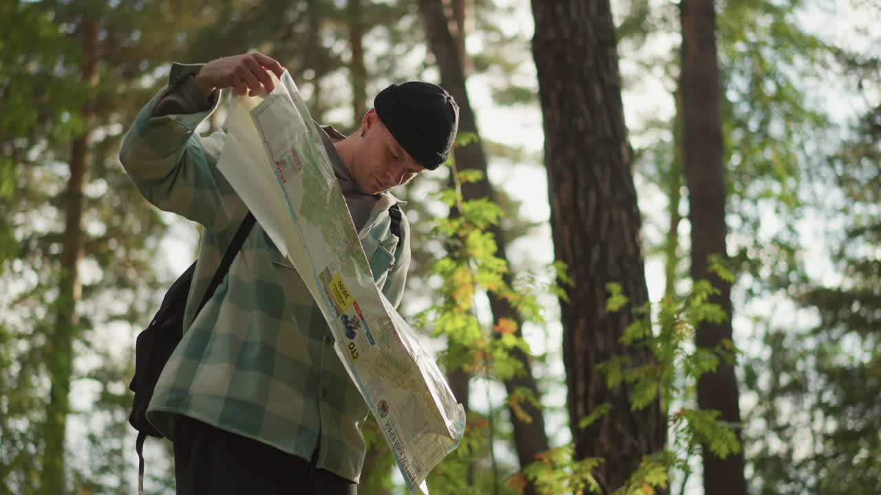 White hiker unfolding large map in forest, hands smoothing creases, focused gaze, scoutlike posture, sunlight filtering through canopy, planning route, map study, trail guide action