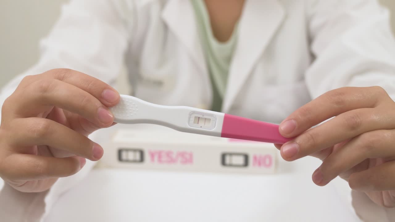 Doctor's hands holding a positive pregnancy test on a white table with the instruction box behind