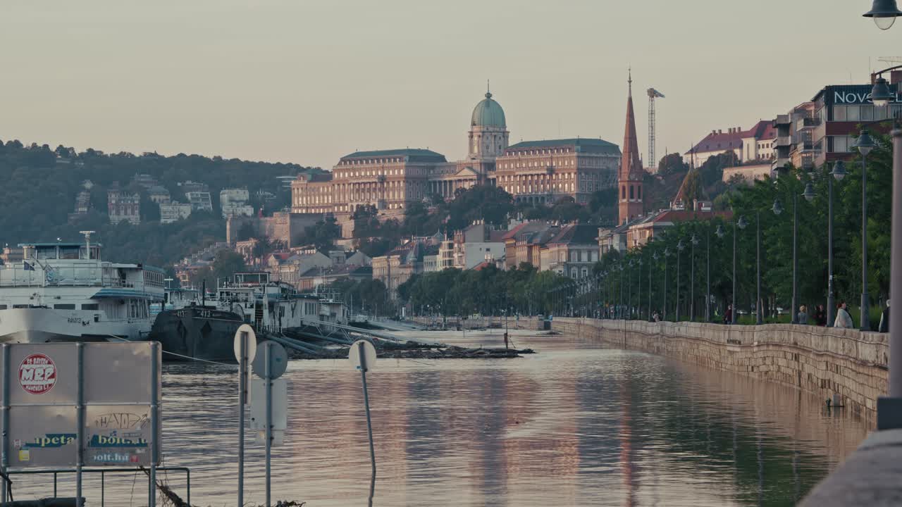 Flooded riverside walkway with traffic signs and boats visible, with the Buda Castle in the background during the Budapest Flood 2024