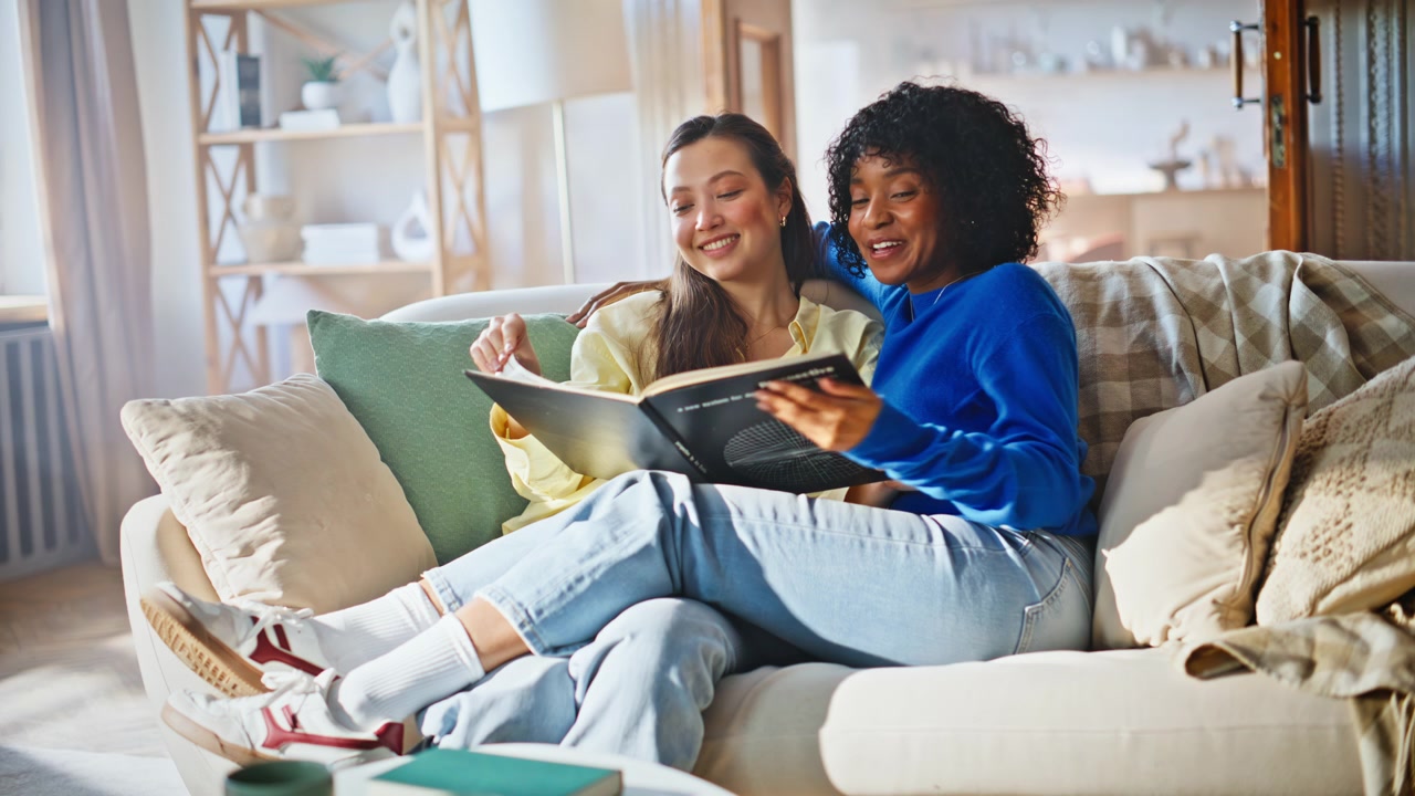 Pareja interracial leyendo un libro en el sofá. Mujeres compartiendo un estilo de vida hogareño y acogedor de fin de semana