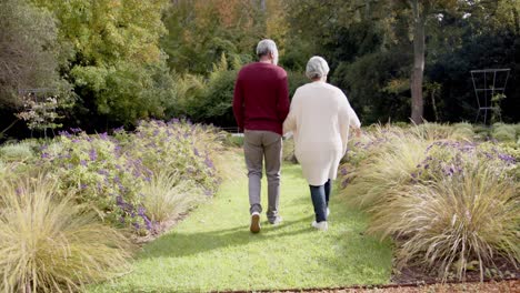 Senior-biracial-couple-holding-hands-and-walking-together-in-sunny-garden,-unaltered,-in-slow-motion