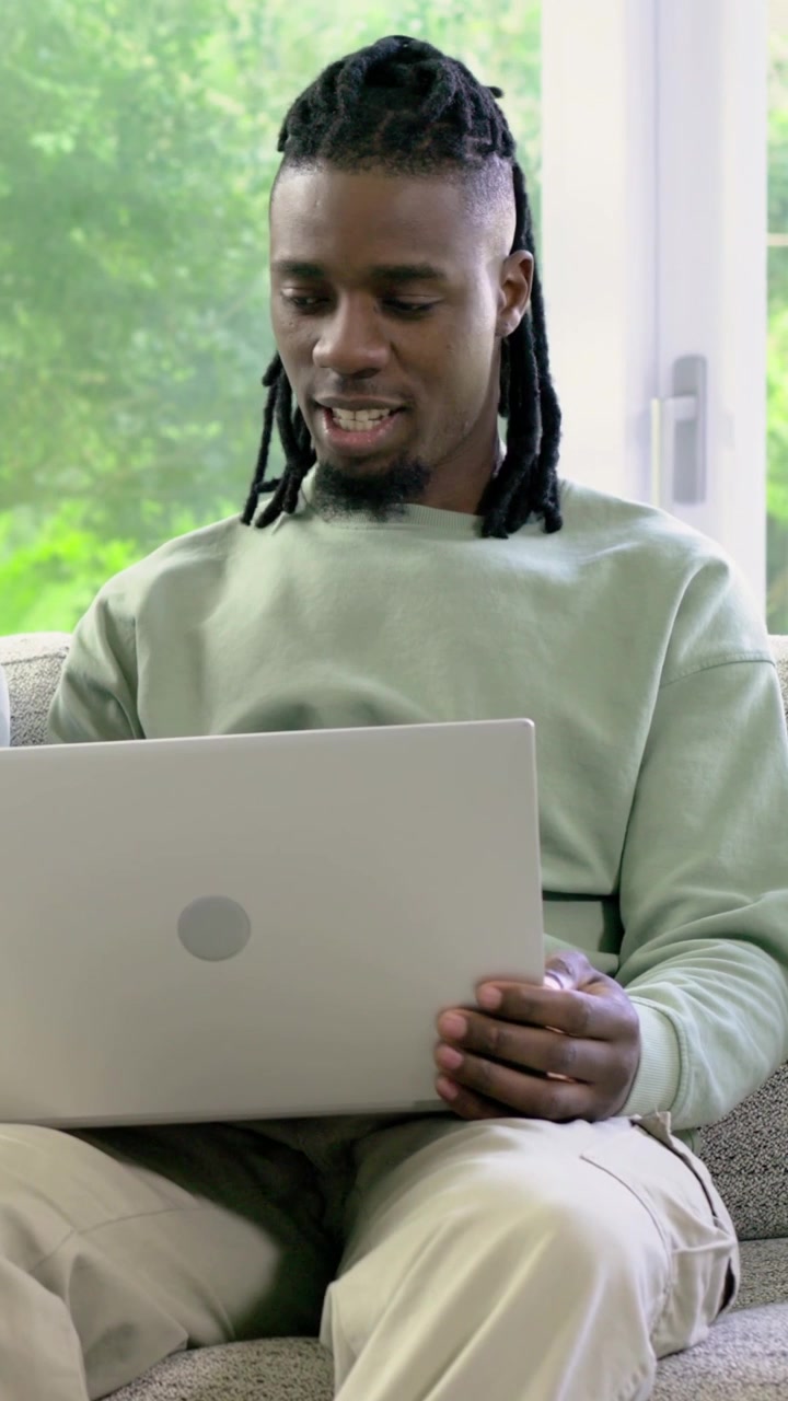 Vertical video: Sitting on sofa at home near window, man opening silver laptop typing and smiling
