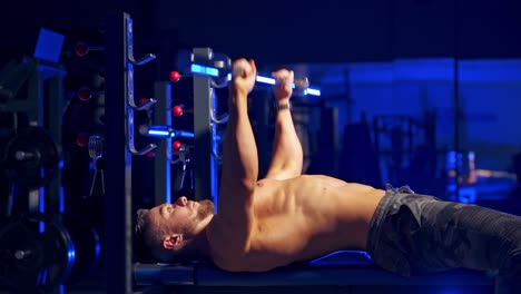 shirtless young bodybuilder doing bench press for chest. strong abs. blue light filter. video from the side.