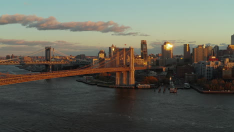 Large-suspended-bridge-lit-by-setting-sun.-Cruise-ship-heading-to-Brooklyn-Bridge.-Buildings-on-waterfront.-Brooklyn,-New-York-City,-USA