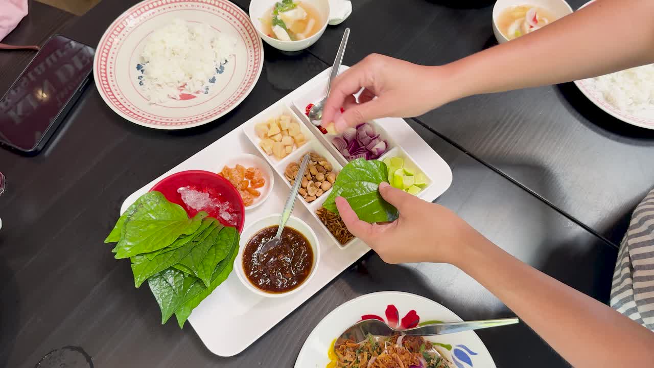 Hands assemble Miang Kham by placing assorted fillings onto green leaves at a well-lit dining table, using overhead camera angles for a clear view