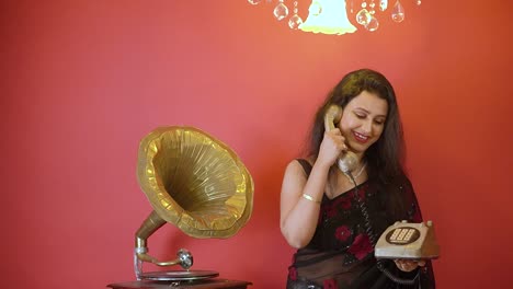 una mujer india con sari negro hablando por teléfono atrapada en la mano, de pie junto a un gramófono en un fondo rojo aislado con luz dorada en la parte superior