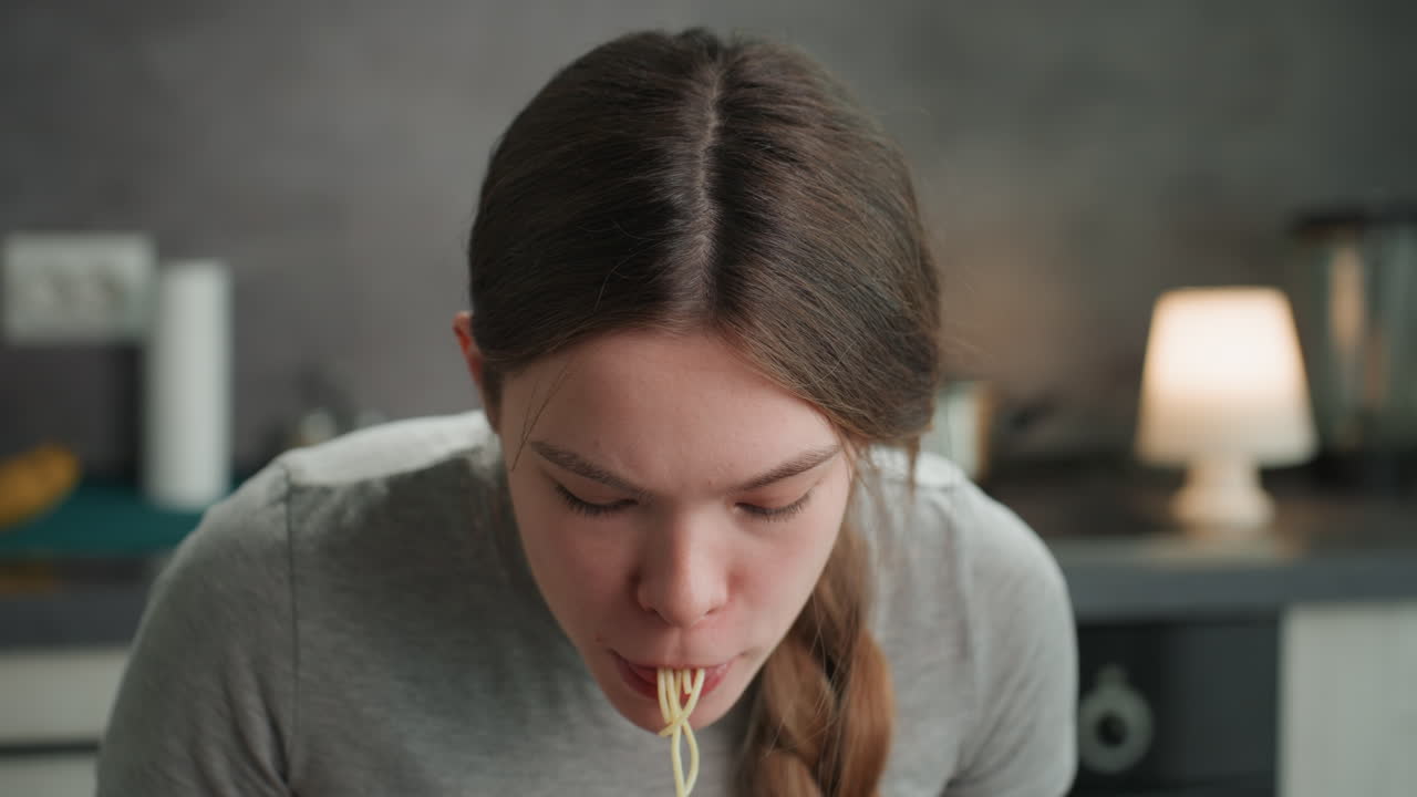 キッチンで麺を食べる妊婦。湯気が立ち上る丼から麺をすすり、唇にはソース。努力と安堵の間で揺れ動く集中した表情、編み込みの髪、そして居心地の良い家庭的な照明。