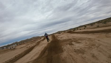 Siguiendo-Una-Moto-De-Motocross-Todoterreno-Que-Da-Un-Salto-En-Una-Pista-De-Carreras-Con-El-Telón-De-Fondo-Del-Desierto-De-Mojave---Drone-Aéreo-En-Primera-Persona-En-Cámara-Lenta