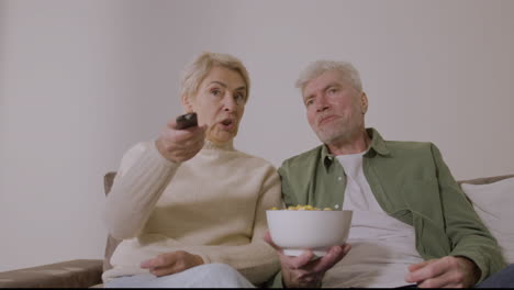 happy senior couple eating caramel popcorn and watching tv while sitting on sofa at home