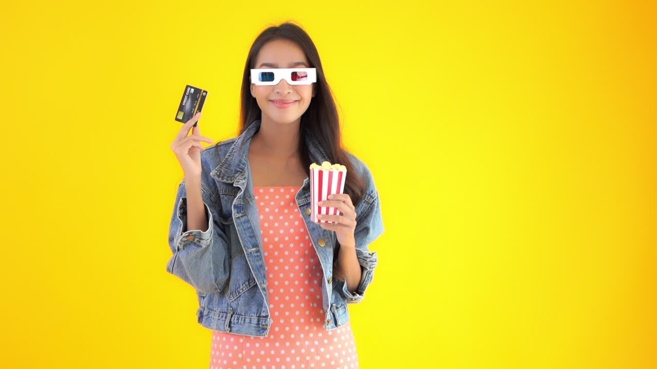 A young pretty woman holds a credit card in one hand, a box of popcorn in another while wearing 3D glasses