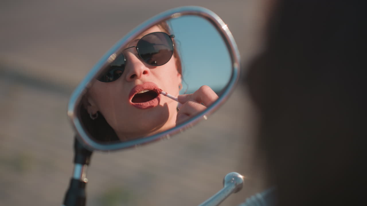 Primo piano di una donna chic con occhiali da sole che si mette il rossetto usando lo specchietto laterale di una moto alla luce del sole, mettendo in risalto bellezza, eleganza e uno stile di vita all'aria aperta sicuro di sé