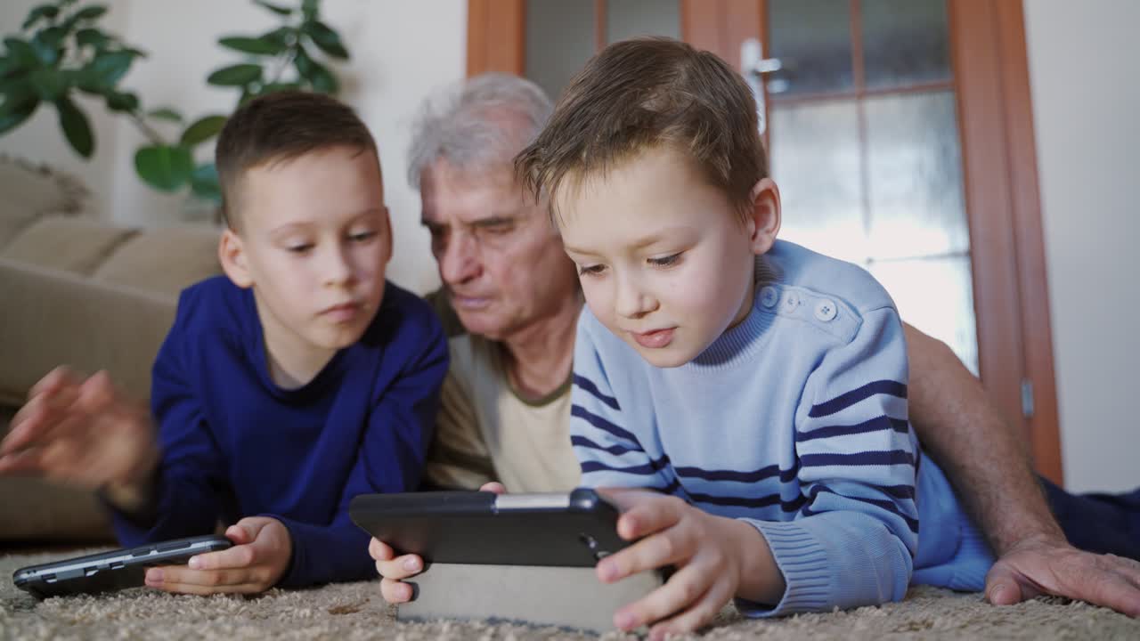 Two boys are playing on the tablets under the supervision of a grandfather in the room on the background of sofa and green plant on the weekday. Relatives.