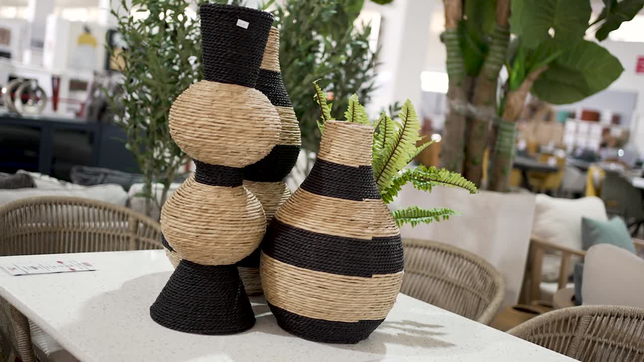 Beautifully crafted woven vases in unique shapes with a mix of black and natural fibers sit on a table in a home decor store. The bright space features stylish furnishings and greenery