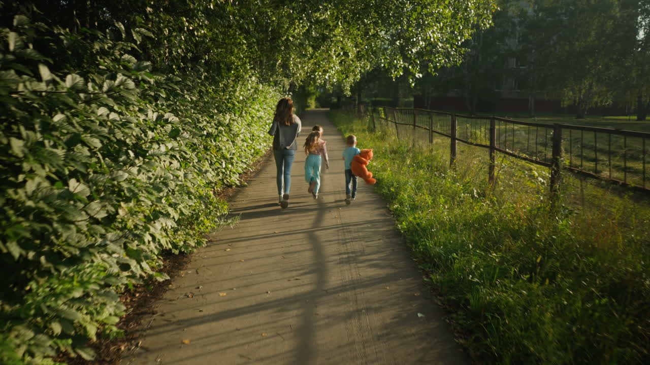 Rear view of cheerful woman and children playfully running together along quiet paved path surrounded by lush greenery, boy carrying teddy bear, enjoying carefree family bonding under warm sunlight