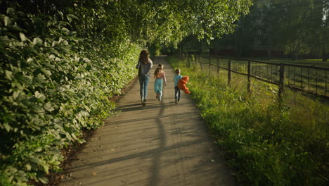 rear view of cheerful woman and children playfully running together along quiet paved path surrounded by lush greenery, boy carrying teddy bear, enjoying carefree family bonding under warm sunlight