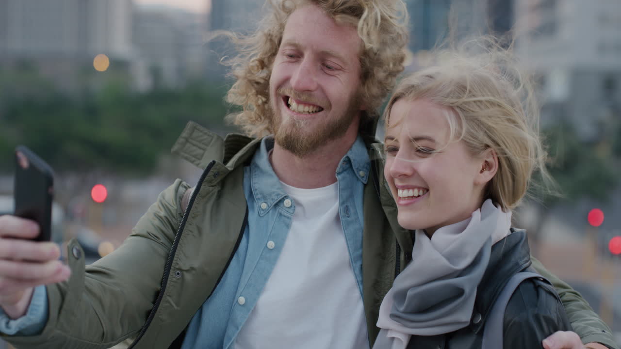 portrait happy caucasian couple using smartphone smiling taking selfie photo enjoying windy day in city sharing urban travel experience on mobile phone camera technology slow motion