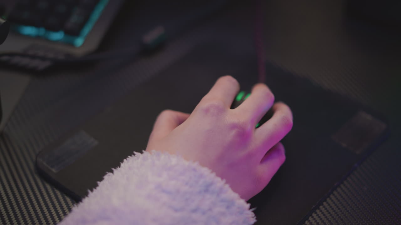Close up of hand wearing fluffy sweater using mouse on dark mouse pad, illuminated by ambient blue and pink lighting, keyboard slightly visible in background
