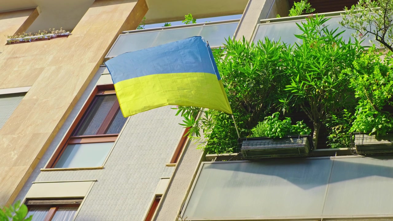 Una bandiera ucraina blu e gialla esposta da un balcone circondato da piante rigogliose su un moderno edificio residenziale a Milano, Italia
