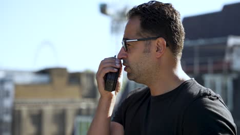 hombre con gafas y cuerpo atlético usando un walkie talkie en un techo en un día soleado
